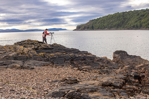 A middle aged female dressed in a red and white waterproof and peaked cap is using a bird watching telescope on a tripod. She is standing on some coastal rocks with the sea (Loch Torridon) and distant hills behind. The area is Lower Diabaig on the western coast of Scotland.