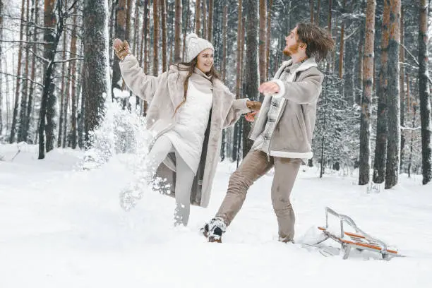 Love romantic young couple girl,guy in snowy winter forest sledding,playing.Walking with sleigh in stylish clothes, fur coat,jacket, woolen shawl,bonnet.Snow lovestory.Romantic date,weekend.