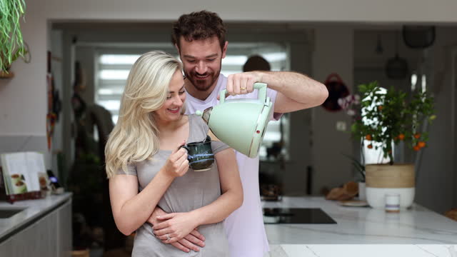 Loving man hugging his partner from behind serving water from the kettle and kissing her very playfully and smiling