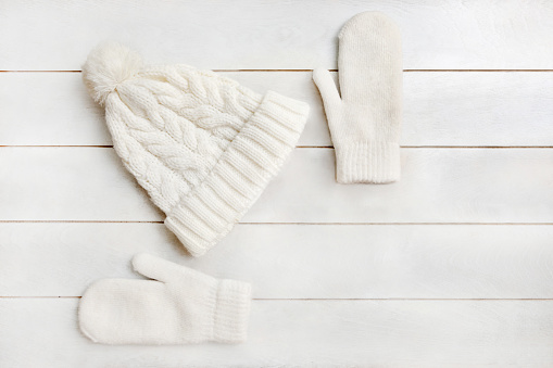 A white knitted hat with a pompom and woolen mittens lie on a white wooden background. Details of winter clothing, flat lay, top view