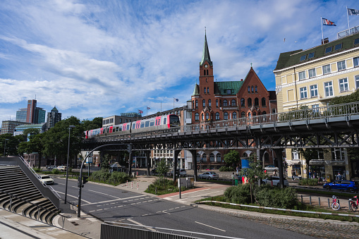 This horizontal photograph offers a layered perspective of Hamburg's bustling harbor area. Starting from the promenade, a set of stairs descends to the street level where pedestrians are seen walking. Above the street, the metro track adds another layer to the urban landscape. In the distance, various buildings and moving cars contribute to the city's dynamic backdrop. All under a blue sky dappled with clouds, captured on a sunny summer afternoon.