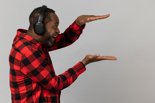 Delighted black bearded man in headphones wearing red checkered shirt showing advertisement area posing isolated over gray studio background.