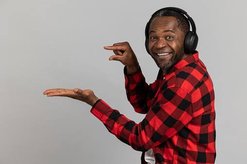 Cheerful black bearded man in headphones wearing red checkered shirt showing small size and advertisement area on palm posing isolated over gray studio background.