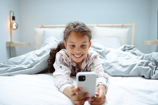 Cute girl in pajamas laying down in bed and smiling while using smart phone