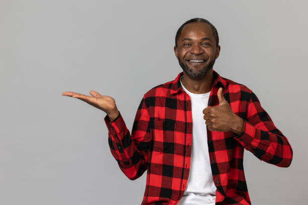 istock Joyful black bearded man wearing red checkered shirt presenting copy space on palm showing thumb up 1689151674