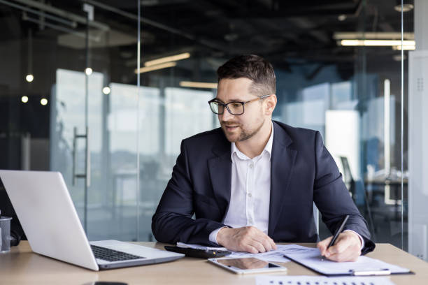 istock Concentrated young businessman office worker working at workplace with documents. Looks at the laptop monitor and takes notes 1689159424