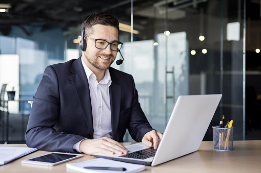 A young businessman is talking in the office on the news of a call from a laptop. He sits in the office in a headset, consults, supports, advises.