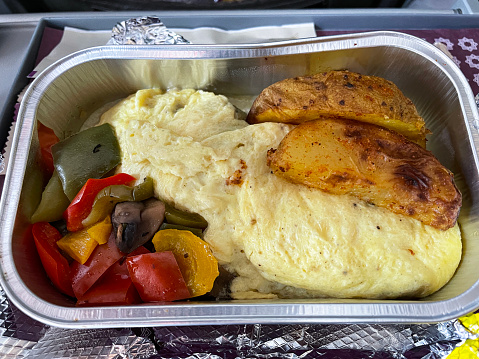 Stock photo showing an airplane flip down table with foil food tray containing a breakfast of omelette, potato wedges and chopped red, green and yellow peppers.