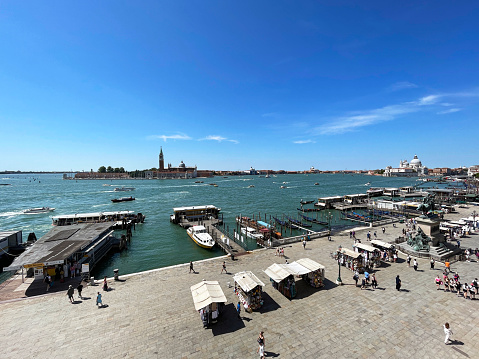 Venice, Italy - July 14, 2023: Stock photo showing close-up view of  tourists and locals visiting the market stalls on Riva degli Schiavoni with moored boats at Venice lagoon waterfront wooden jetties with a background view of San Giorgio Maggiore, Venice, Italy.