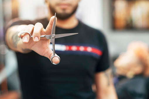 Stylist with a beard on a black T-shirt holding scissors in his hand stretched out towards the camera. Selective focus on the scissors