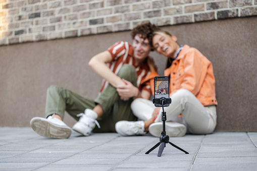 Portrait of a cheerful heterosexual couple sitting on the concrete floor and posing to the smart phone camera. The smart phone is fixed to the tripod.