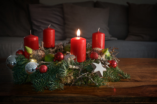 Advent wreath with one burning red candle and Christmas decoration on a wooden table in front of the couch, festive home decor for the first Sunday, copy space, selected focus, narrow depth of field