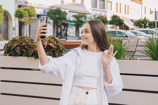 Beautiful european girl with long hair in total white look on the street talks by videocall and articulates with her hand. Banner, place for text concept.