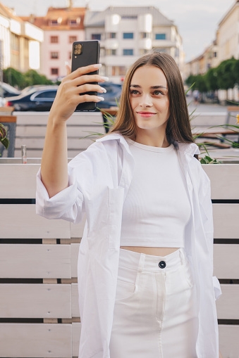 Beautiful european girl with long hair in total white look on the street filming a video or has a videocall. Vertical photo, social networks concept.