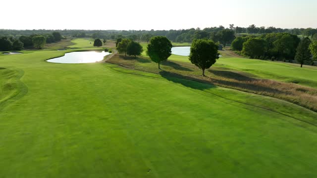 Prestigious green golf course in USA. Aerial shot of long fairway with pond water feature reflecting golden light during sunset.