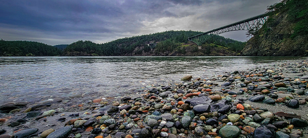 Colorful pebbles line a riverbank with a high bridge in the background