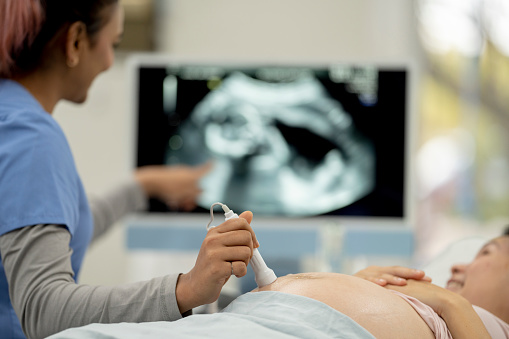 A pregnant mother lays out on an exam table with her belly exposed, as a female sonograhpher takes a look at her baby with an ultrasound machine.  The patient looks at the screen to see her baby as the technician point out the various parts of the baby.