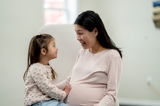 A little girl places her hand on her Mothers belly as she tries to feel her new sibling moving.  She is dressed casually and looking up at her mother with excitement.