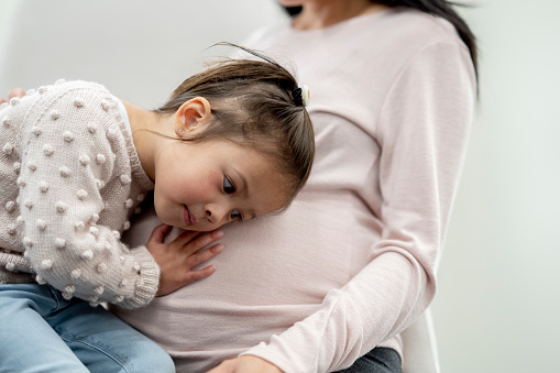 A little girl places her ear on her Mothers belly as she tries to hear her new sibling moving around.  She is dressed casually and has a neutral expression on her face.