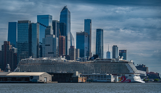 A cruise ship awaits to depart in a New York pier on a clear day. The modern Midtown skyscrapers tower over the massive ship while at port.