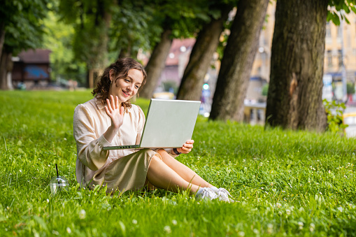 Portrait of pretty young woman making video laptop webcam conference call with friends or family enjoying pleasant conversation, waving hello, laughing. Girl sitting on grass in urban sunset city park