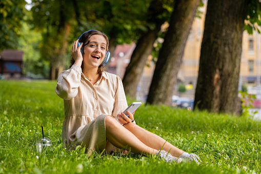 Happy relaxed overjoyed young woman in wireless headphones choosing, listening favorite energetic disco rock n roll music in smartphone dancing outdoor. Girl sitting on grass in urban sunset city park