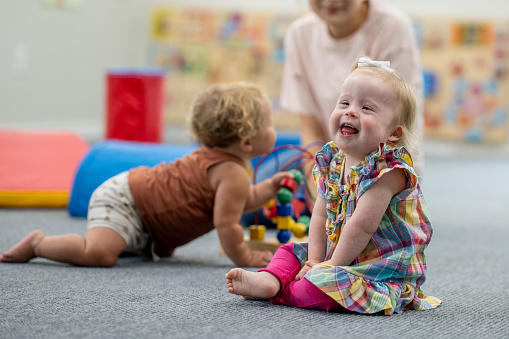 A sweet little girl with Down Syndrome plays on the floor at daycare with a little friend and her Childcare provider.  She is dressed casually and has a smile on her face as she giggles joyfully.