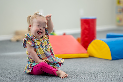 A sweet little girl with Down Syndrome sits on the floor as she plays with large colourful foam blocks at Daycare.  She is dressed casually and has a smile on her face as she giggles.