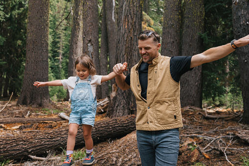 Caucasian father holds preschool aged Eurasian daughter's hand, helping her walk across a fallen tree trunk while spending time together hiking in Oregon.