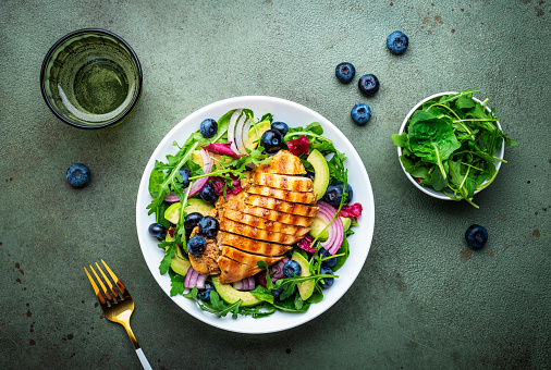 Delicious salad with blueberries, grilled chicken, avocado, red onion, walnuts, arugula and mixed herbs, green table background, top view
