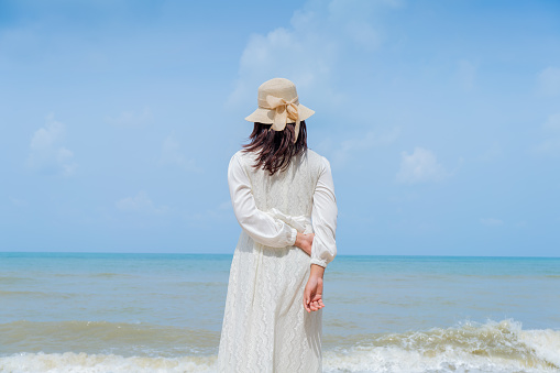 Happy Young woman in a white dress and hat standing on the beach and enjoying freedom, and looking at the sea.