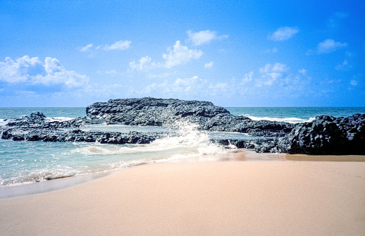 Vintage 1980s film photograph of the lava fields of black volcano rock against the blue waters and sandy beach on the tropical island of Maui, Hawaii.