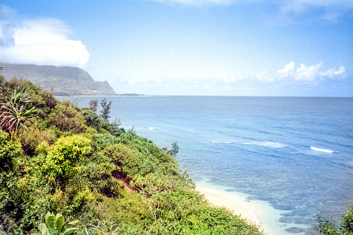 A vintage 1980s film photograph view perched on an ocean bluff overlooking Hanalei Bay, Pali Ke Kua Kauai, Hawaiian volcanic coastline with hidden beach tucked in the coastline.