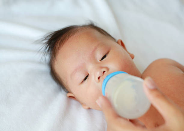 silicone baby bottle with a nipple, half-filled with milk, rests on a bed. In the background, a silhouette of a baby can be seen, creating a nurturing and cozy atmosphere
