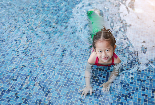 Smiling little Asian child girl in a mermaid suit playing poolside with looking camera.