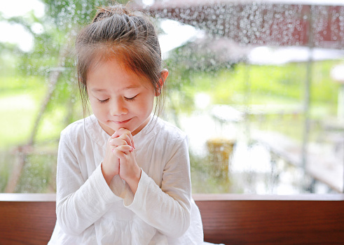 Adorable little Asian girl praying at glass windows on the raining day. Spirituality and religion.
