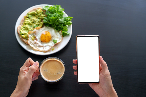 A woman's hand holding a smartphone while having breakfast at home, with the smartphone displaying a blank screen for design mockup