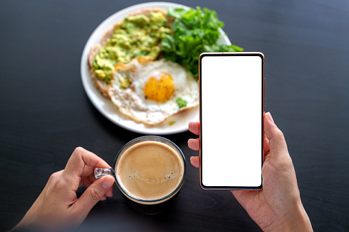 A woman's hand holding a smartphone while having breakfast at home, with the smartphone displaying a blank screen for design mockup