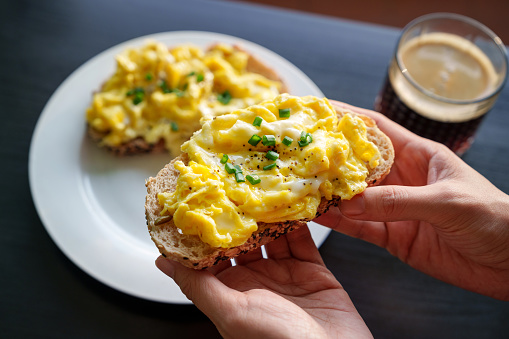 A cropped image of a woman's hand holding a piece of toasted bread with scrambled eggs on top, as she enjoys a healthy breakfast in the morning.