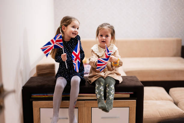istock Two sisters are sitting on a couch at home with british flags on hands. Great Britain children girls with flag . 1690140235