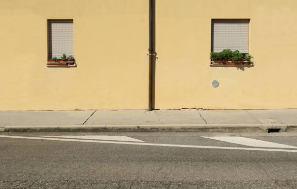 istock Brown roof gutter on yellow plaster wall with two closed windows by sides. Concrete sidewalk and street in front. 1690155975