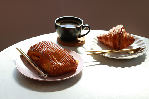 French pastries with a cup of hot black coffee are placed on a wooden table next to a window