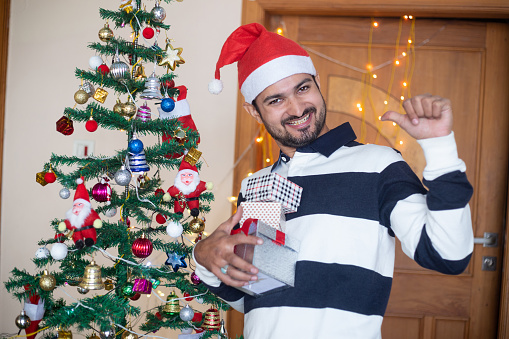 Cheerful Indian Man Wearing Santa hat Holding Christmas Gift Boxes Or Presents Enjoys Holiday Season At Home, Christmas Tree Background
