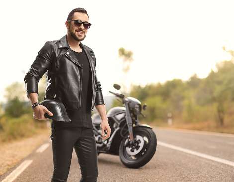 Biker in a leather jacket with parked motorcycle on the road holding helmet and wearing sunglasses