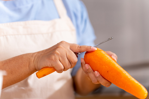 Asian senior woman hands using peeler peeling carrot on kitchen island counter. Elderly woman preparing organic vegetables for cooking healthy food at home.