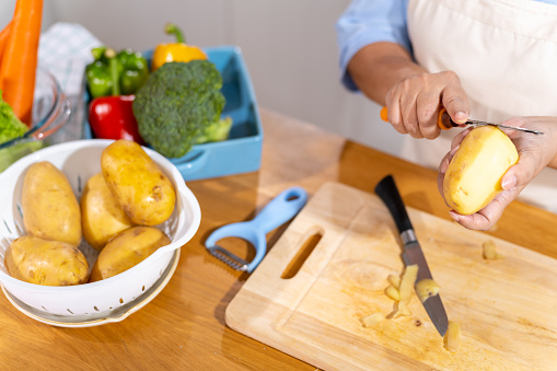 Asian senior woman hands using peeler peeling raw potato on kitchen island counter. Elderly woman preparing organic vegetables for cooking healthy food at home.