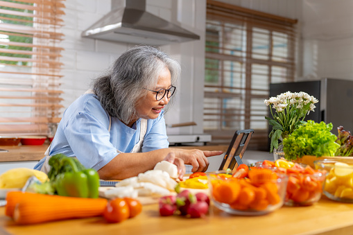 Happy Asian elderly woman following an online recipe on digital tablet with social media on kitchen island counter. Elderly woman enjoy preparing and cooking healthy food in the kitchen for dinner with family at home.