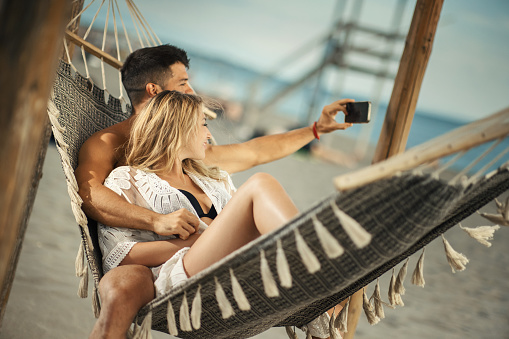 Closeup of a mid 20's couple relaxing at the beach by swinging in a hammock and taking selfies.