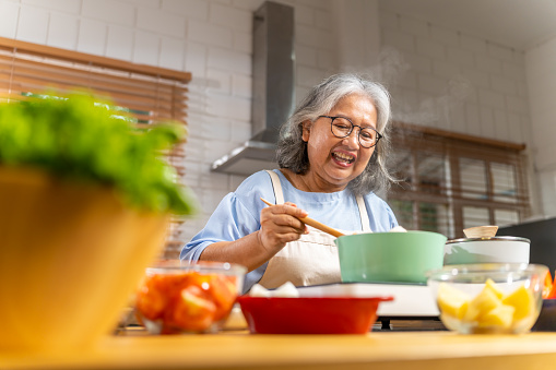 Asian senior woman cooking chicken soup with vegetables on kitchen island counter. Elderly woman enjoy preparing healthy food in the kitchen for dinner with family at home.