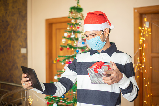 Online christmas celebration, Young man wearing mask congratulating over video calling on tablet showing gift present, covid-19 pandemic, new year, holiday,winter,december,new normal.
